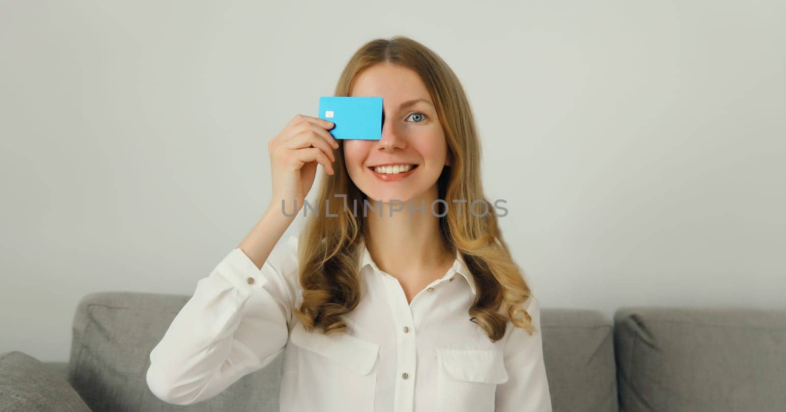 Portrait of happy smiling young woman holding plastic credit bank card, joyful girl customer of a modern bank sitting on the couch in living room at home