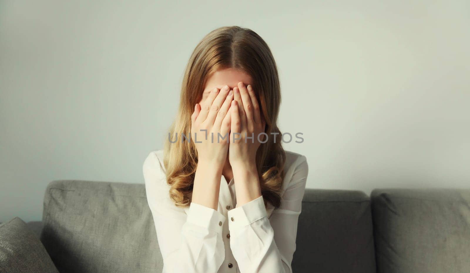Upset sad crying woman covers her eyes and face with her hands while experiencing depression, stress, fatigue, mental suffering sitting on the couch at home