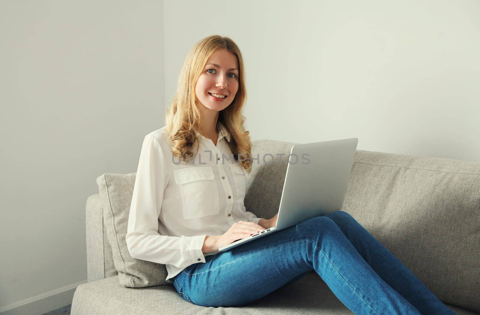 Happy modern young woman working with laptop, freelancer girl smiles looking at camera sitting on a couch in the living room at home