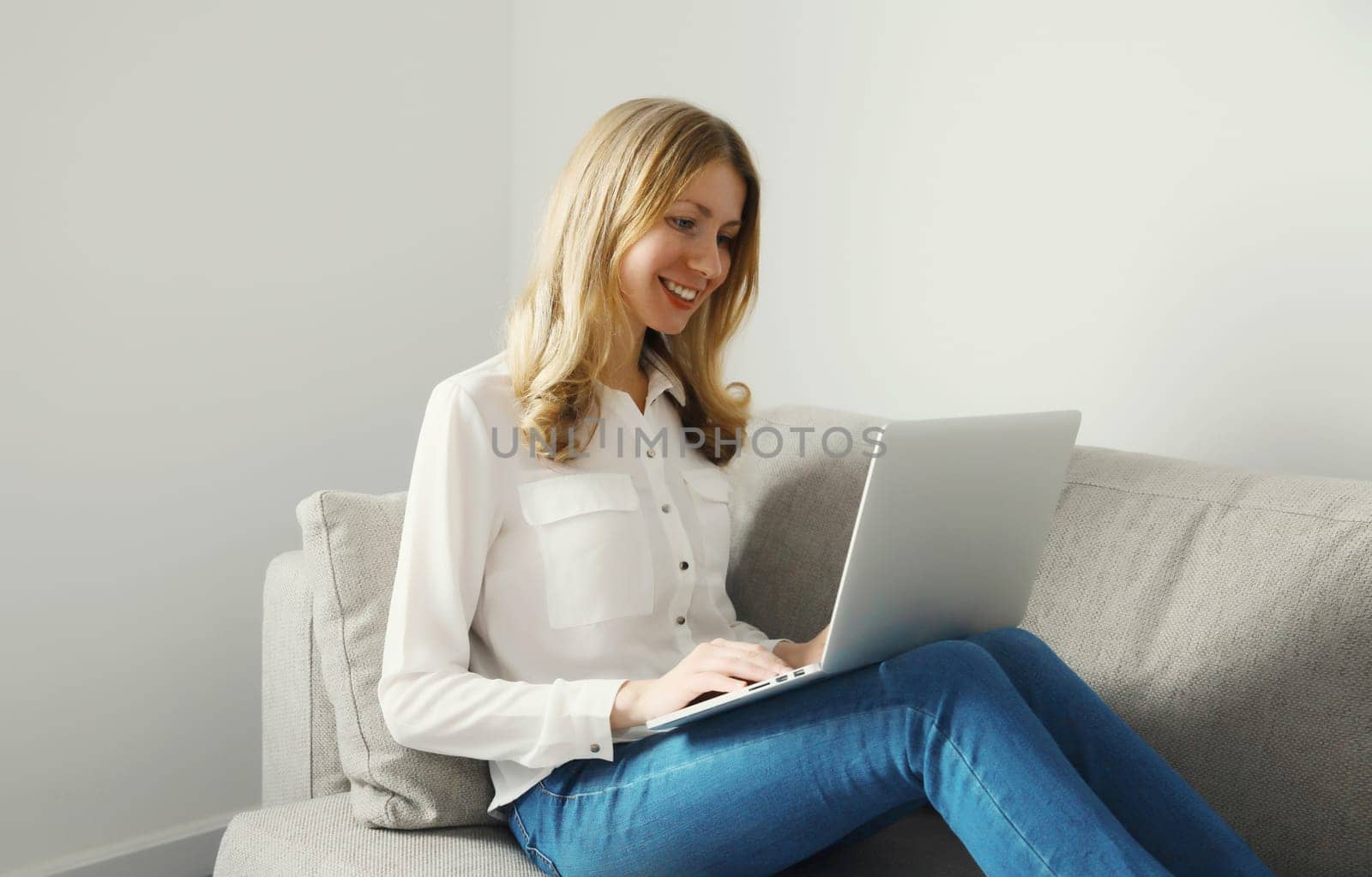 Happy modern young woman working with laptop, freelancer girl smiles looking at computer sitting on a couch in the living room at home