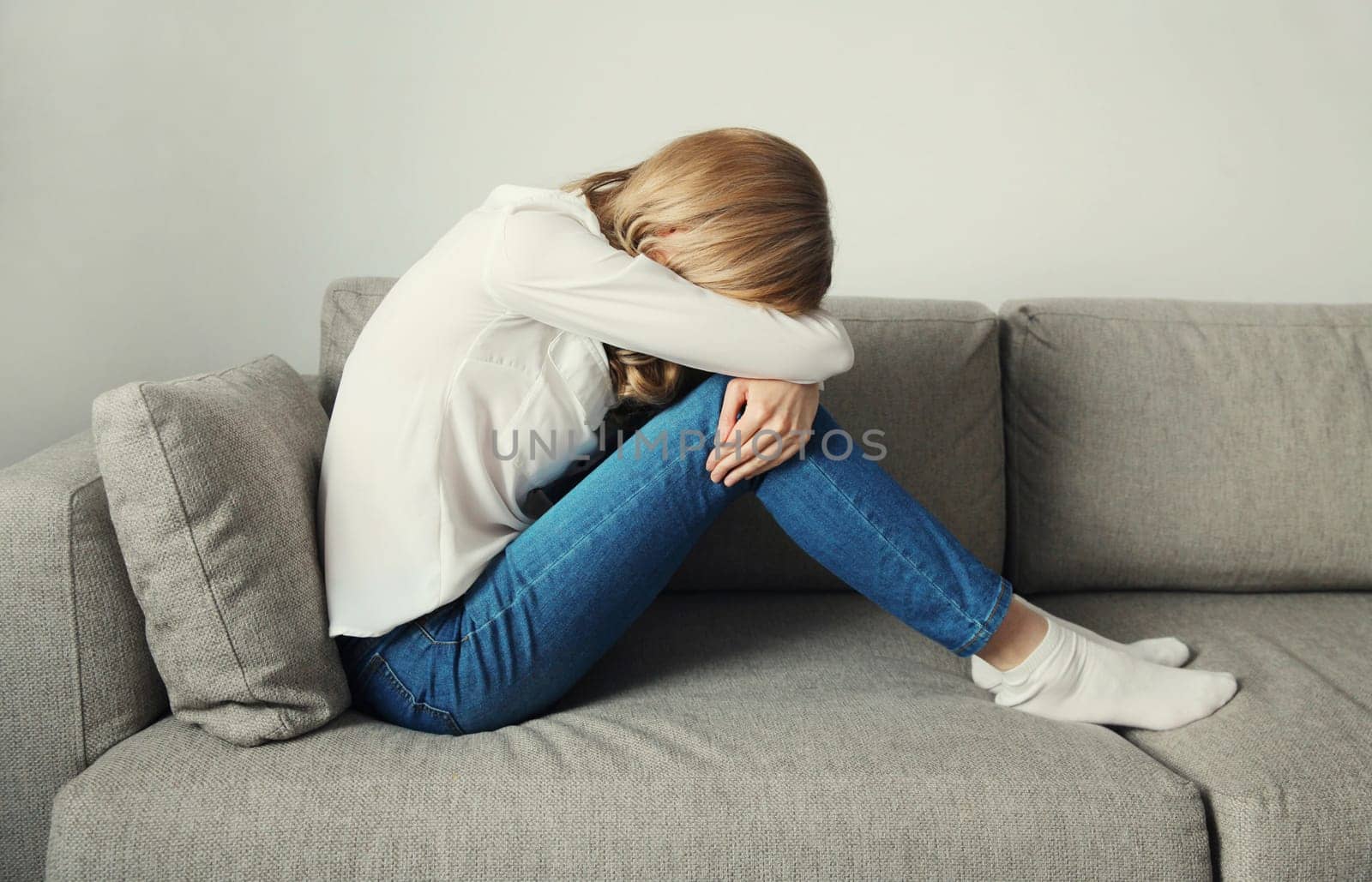Upset sad crying young woman covering her eyes and face with her hands while experiencing depression, stress, fatigue, mental suffering sitting on the couch at home