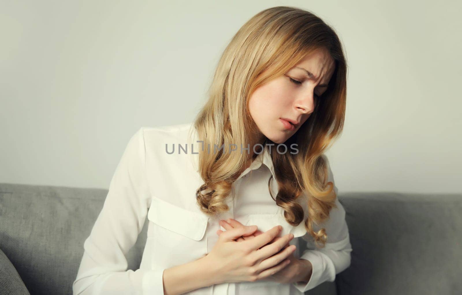 Unhealthy young woman touching hands her chest suffering from heart pain, feeling bad, heart attack, having health problems, sitting on the couch at home