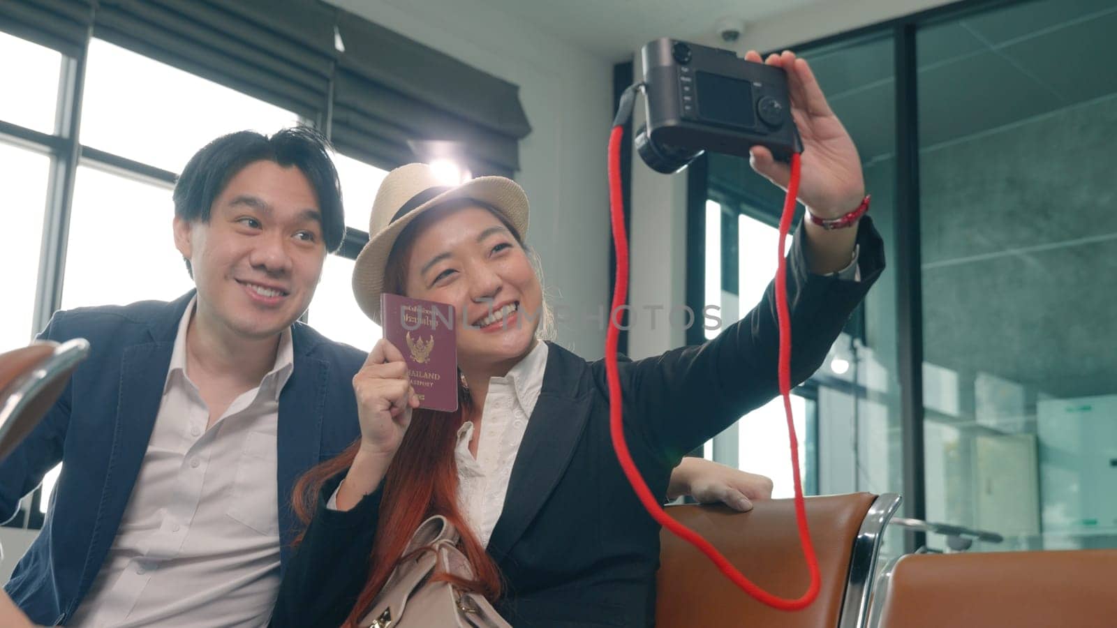 Wait boarding flight. Asian beautiful young loving couple smiling making selfie by their mirrorless camera and holding passports tickets while waiting for boarding in terminal hall airport