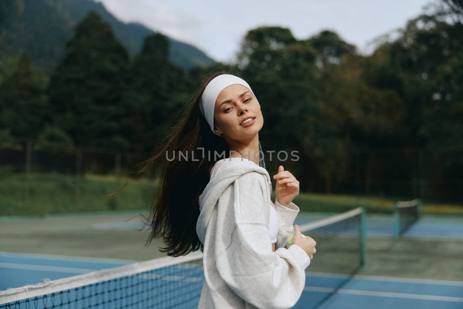 Outdoor tennis practice featuring a young woman in a hoodie and headband, smiling joyfully with her hair flowing, showcasing an active lifestyle in nature