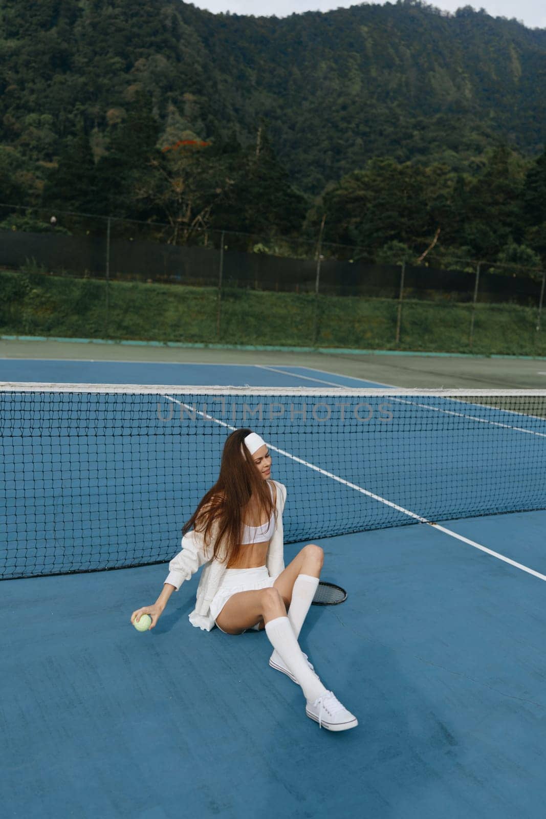 This vibrant scene features a young female athlete in white sportswear, happily resting on a tennis court The lush green mountains create a serene backdrop, perfect for sports enthusiasts