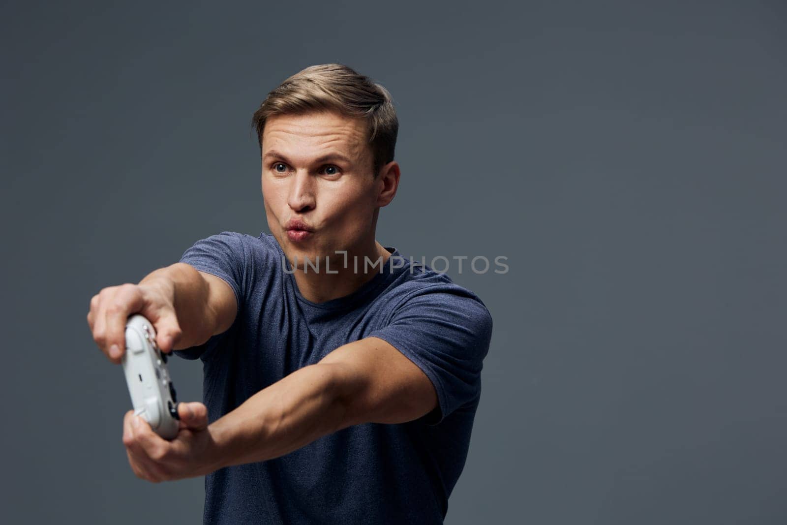 Portrait of a cheerful young man with a fun expression, extending his arms with a game controller in hand, looking engaged and playful, against a simple grey backdrop. Concept: gaming, entertainment, fun activity, casual style, youth.