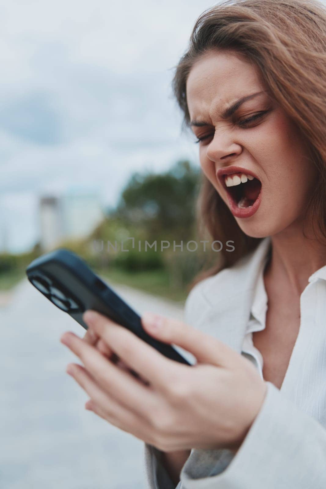 angry woman outdoors holding smartphone with intense frustration and upset expression wearing casual jacket, blurred natural background, concept of financial crisis, stress and emotional struggle