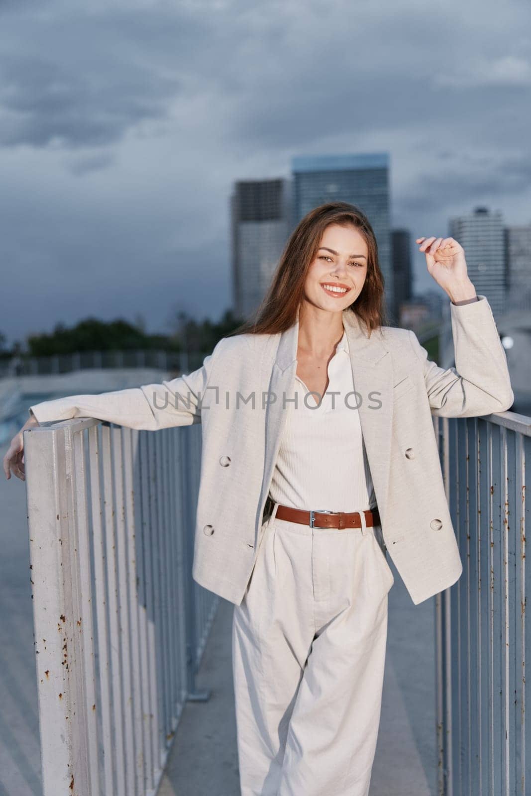 confident smiling woman wearing light beige suit and white blouse stands relaxed on city bridge under cloudy sky, expressing success and modern business fashion style