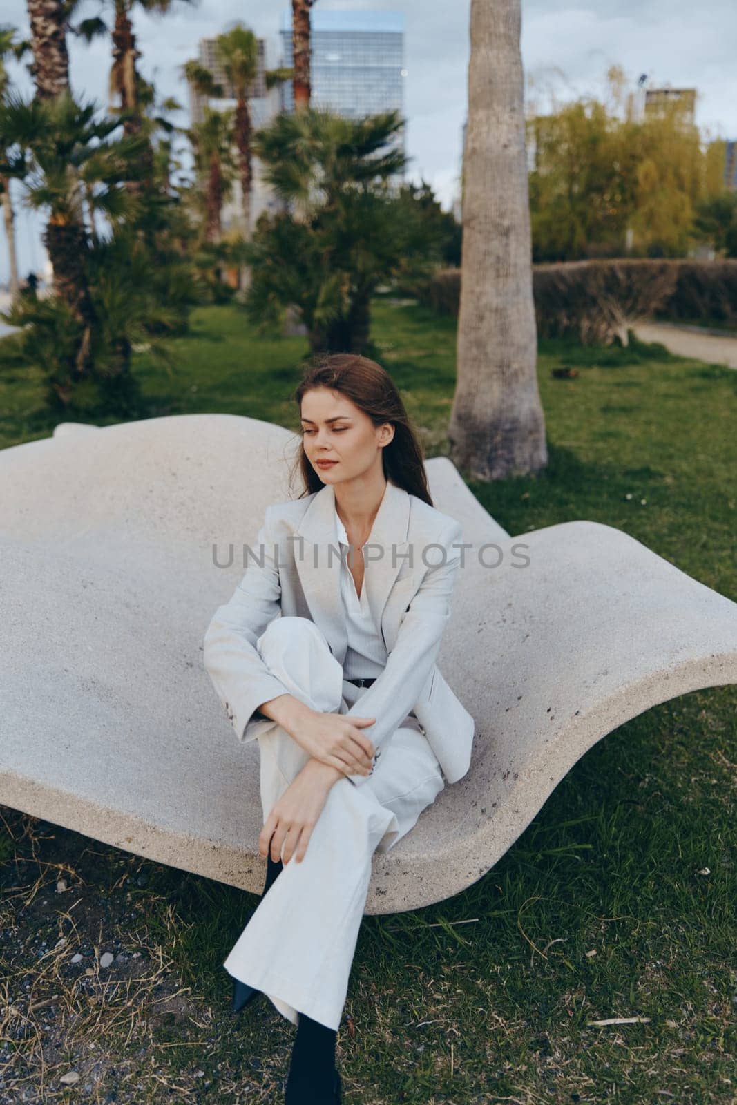 woman in white suit sitting outdoors on modern concrete bench with thoughtful expression, natural green urban park background, business fashion style in spring season