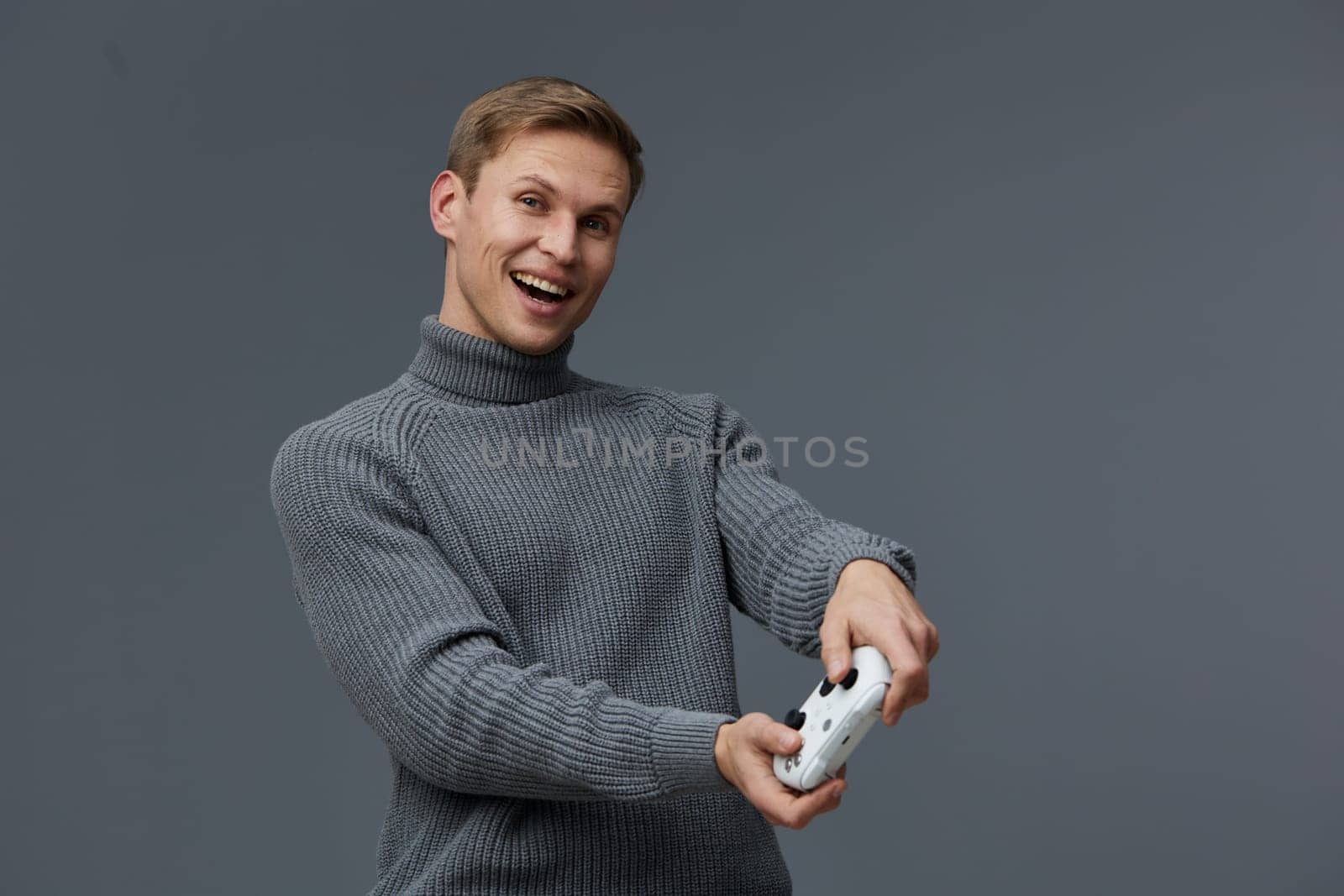 Happy cheerful man with short blond hair in warm gray turtleneck sweater capturing a selfie with smartphone on gray studio background, demonstrating joy and modern technology use. Concept: positive mood and social media