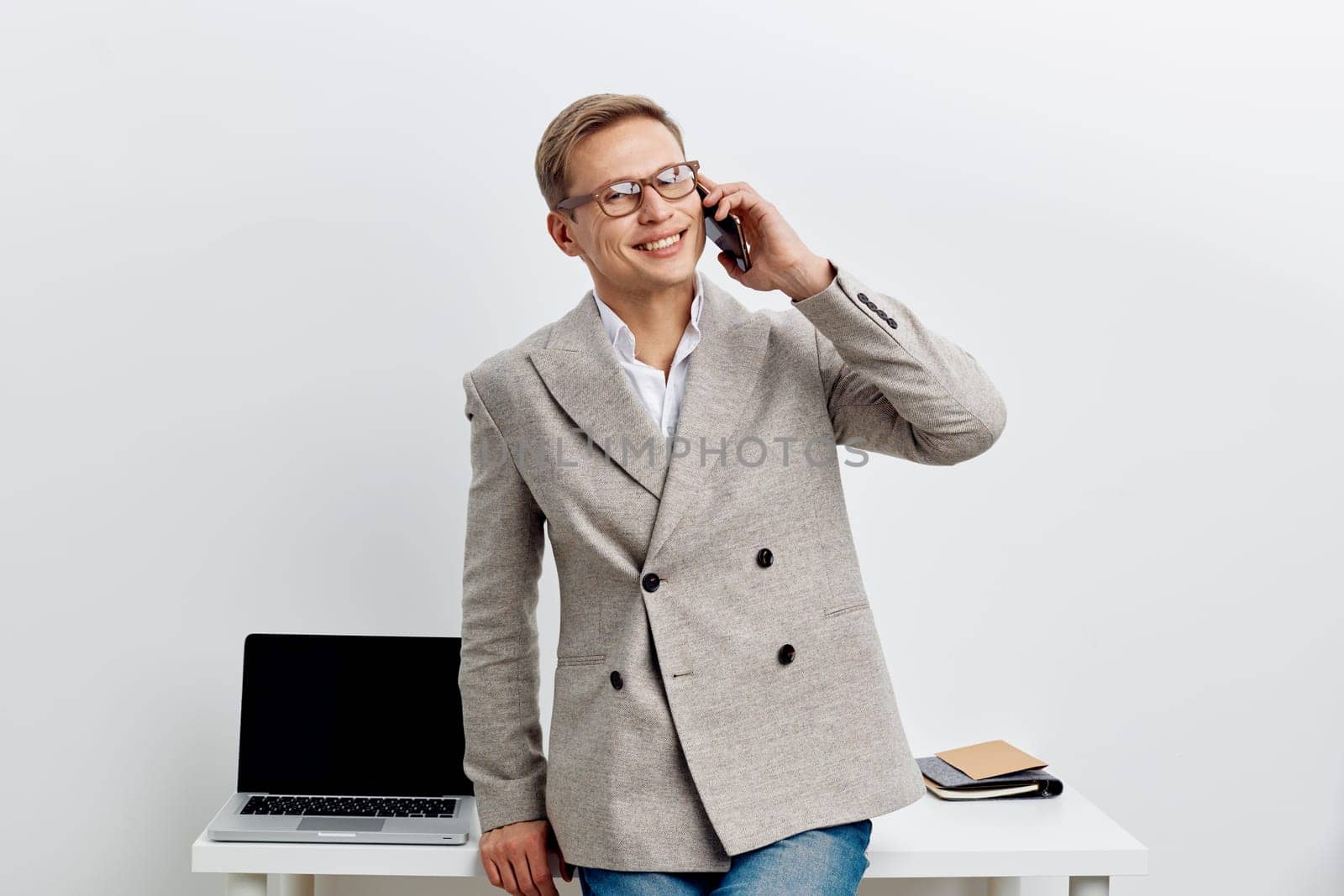 Cheerful man with glasses dressed in gray blazer, holding phone to ear, standing at white desk with open laptop and notebook, modern office background, positive mood, casual business attire. Concept: professional communication