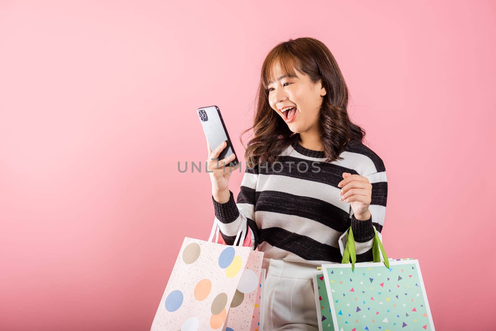 Portrait Asian happy beautiful young woman shopper smiling standing excited holding online shopping bags online multicolor and smartphone on hand, isolated on pink background, glad with mobile phone