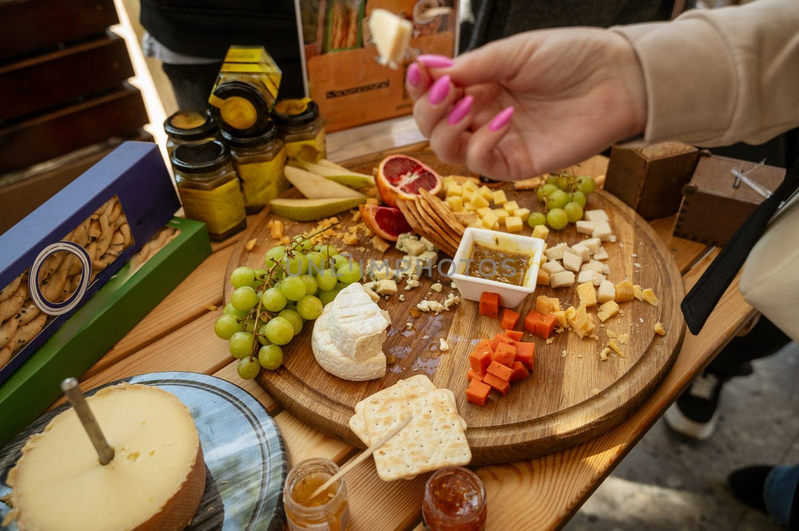 Cheese tasting at a farmers market, close-up of a woman's hand of a buyer. Cheese and fruit platter on the table. Cheese tasting process. Large assortment of natural products on the table. Hand in blur