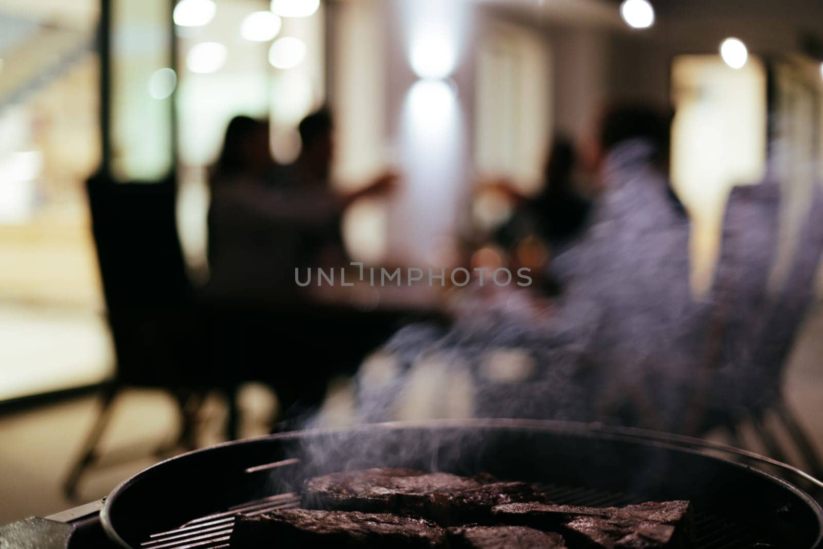 Close-up photo of delicious meat being grilled. In the background, friends and family are sitting and waiting for a meal