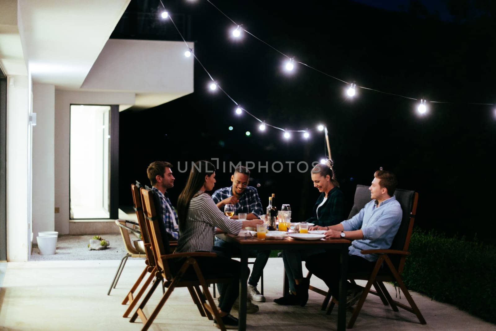 A group of young diverse people having dinner on the terrace of a modern house in the evening. Fun for friends and family. Celebration of holidays, weddings with barbecue.