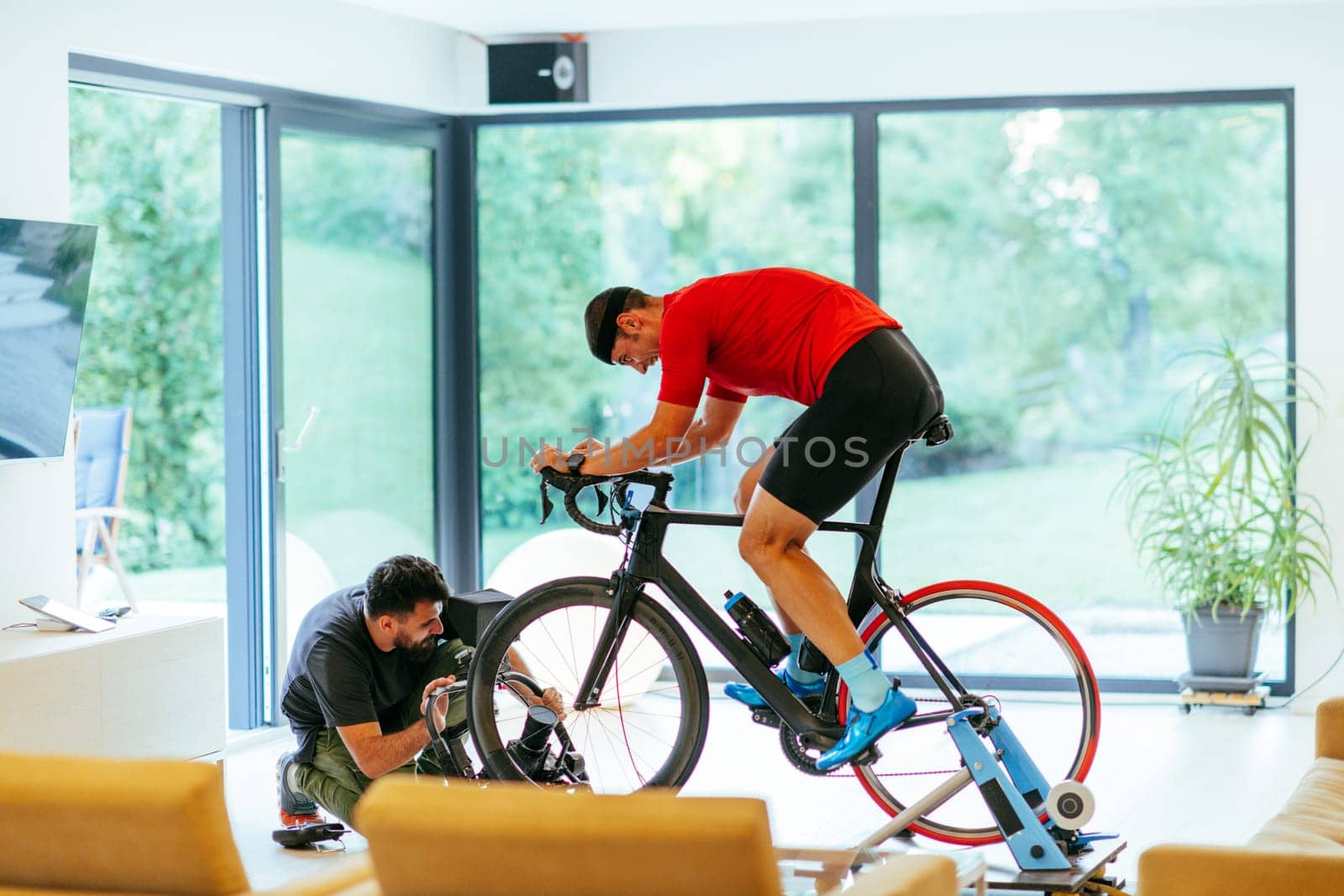 A cameraman filming an athlete riding a triathlon bike on a simulation machine in a modern living room. Training in pandemic conditions