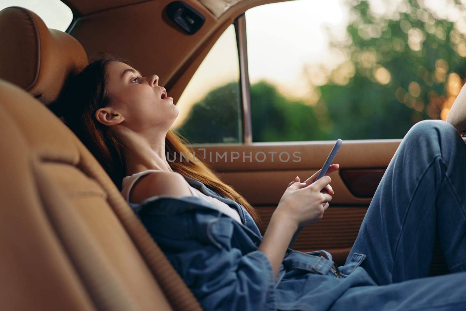 A young woman in casual denim attire, enjoying a moment of solitude in a car, looking at her phone with a serene expression, illuminated by gentle evening light.