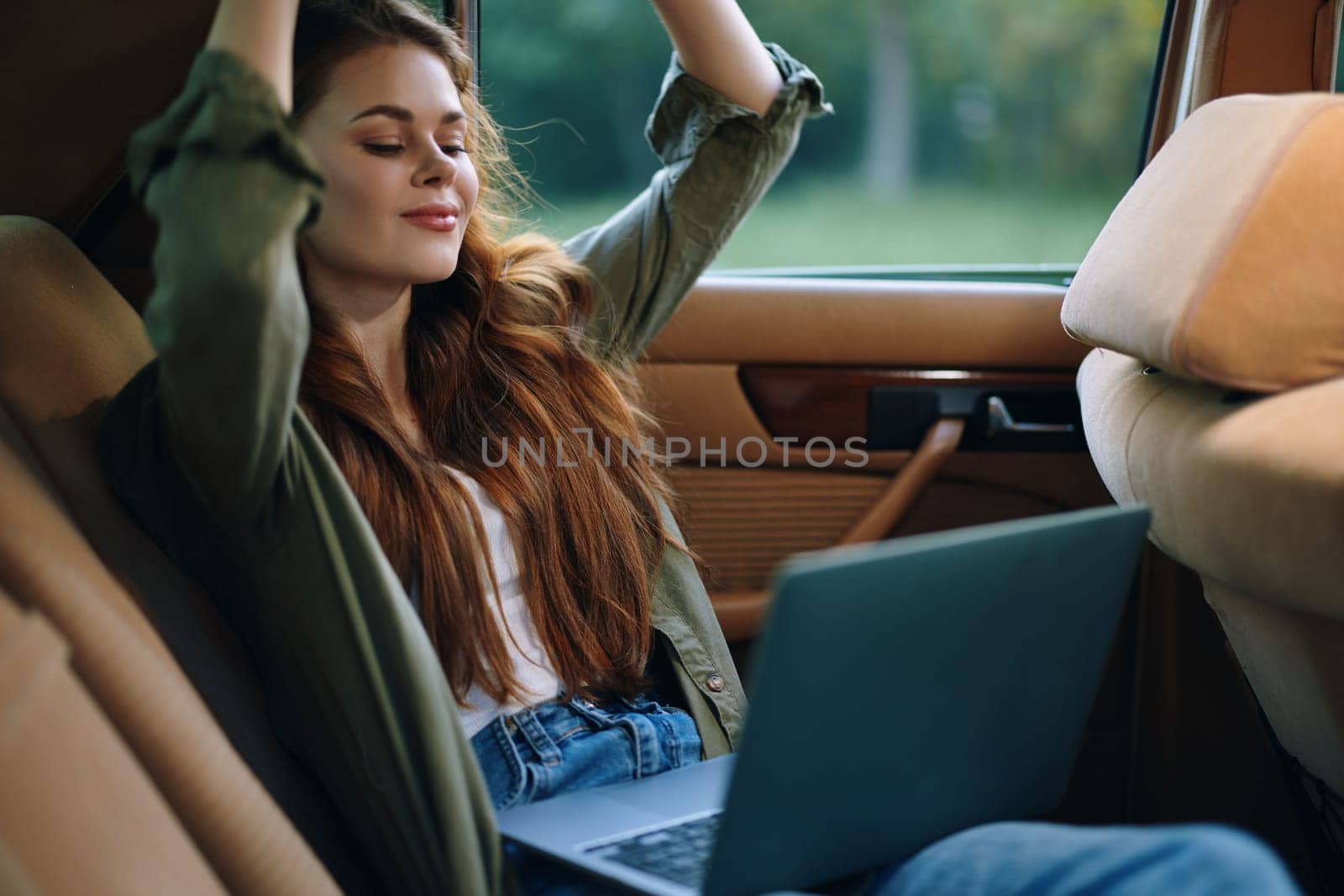 A serene young woman with long hair smiles while using a laptop inside a classic car, dressed in a casual outfit, capturing a moment of outdoor relaxation and work.