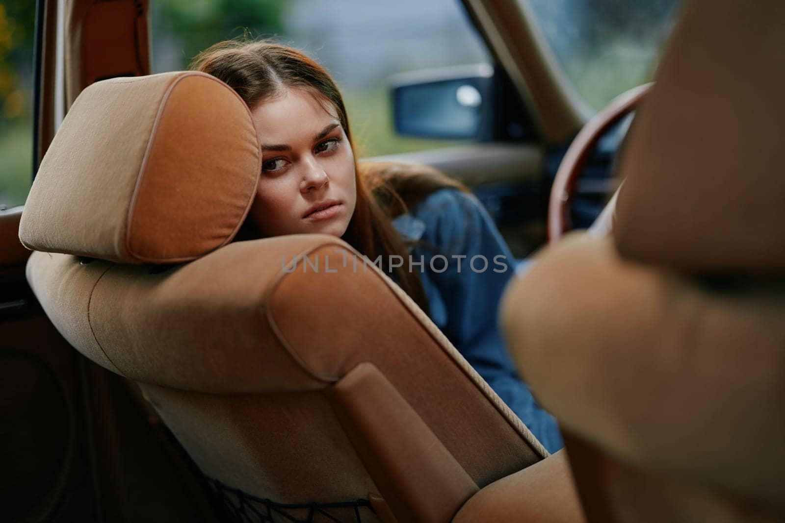 A young woman, dressed casually, appears contemplative and sad as she leans against the seat of a vintage car. The soft color palette and blurred background evoke nostalgia and introspection, making
