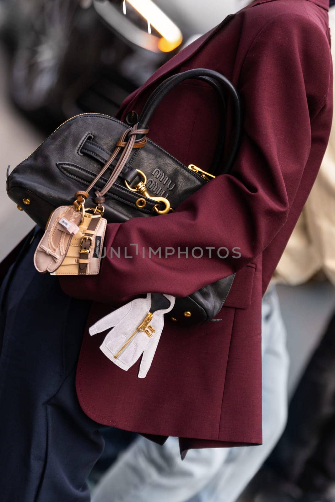 Paris, France - March 11, 2025: Woman wears Miu Miu handbag in black leather with gold rings and mini charm keychains. Worn with wine-red blazer. Street style outfit details.