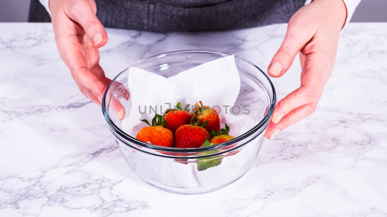 Strawberries, freshly washed and dried, are neatly stored in a glass bowl lined with a paper towel and securely covered with plastic wrap to maintain freshness.