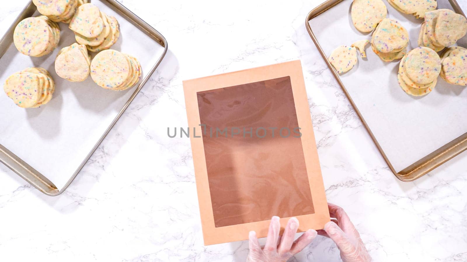 Flat lay. With precision, the woman is carefully arranging the sugar cookies, filled with dough-mixed sprinkles, into a rustic brown paper box.