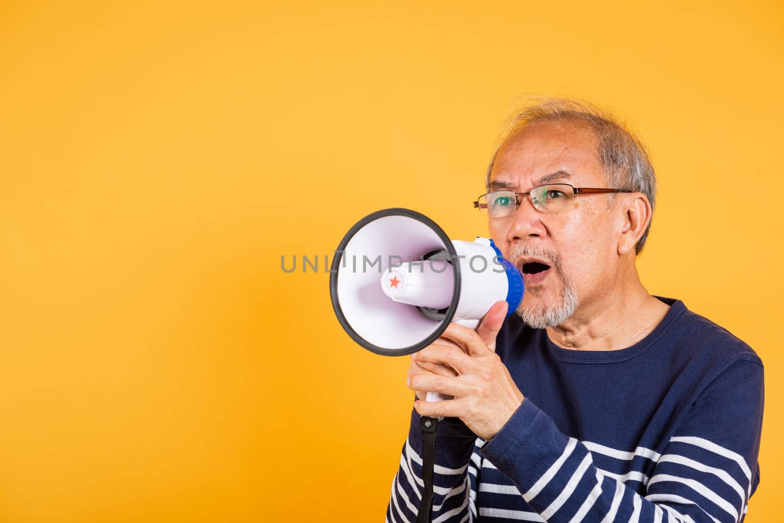 Portrait Asian old man wearing glasses screaming in megaphone studio shot isolated yellow background, happy elderly man holding a megaphone and shouts yelling announces news information