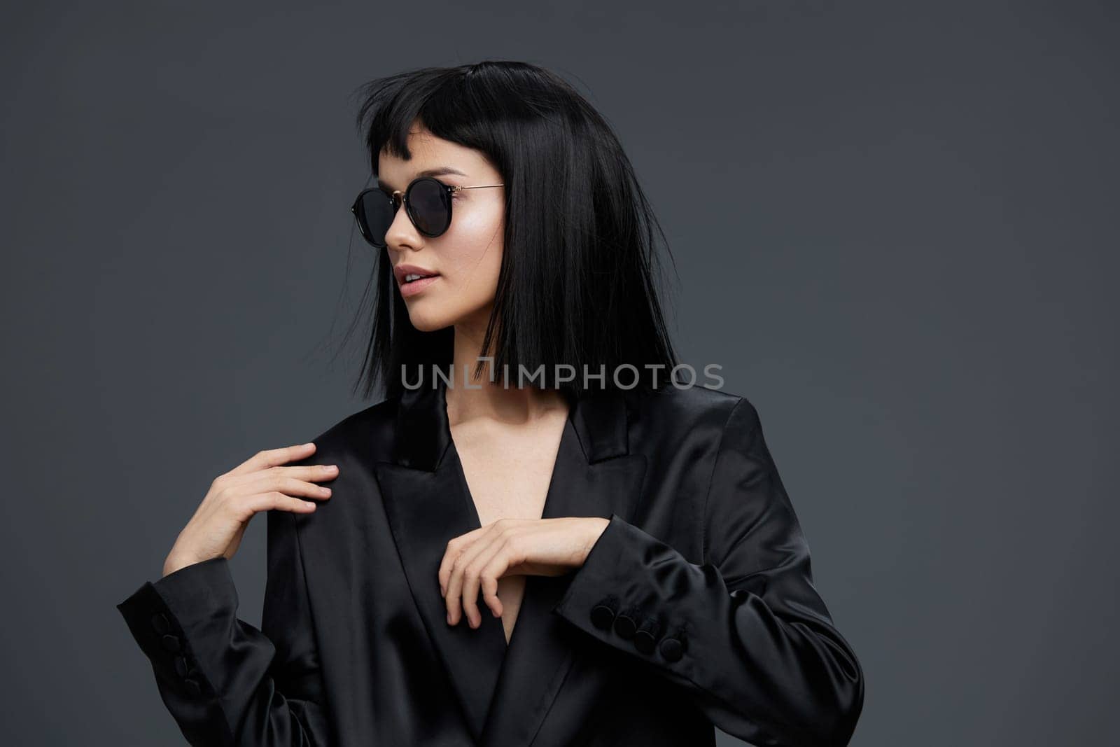 Asian woman in a black suit and sunglasses poses confidently against a gray background, exuding sophistication and modern style