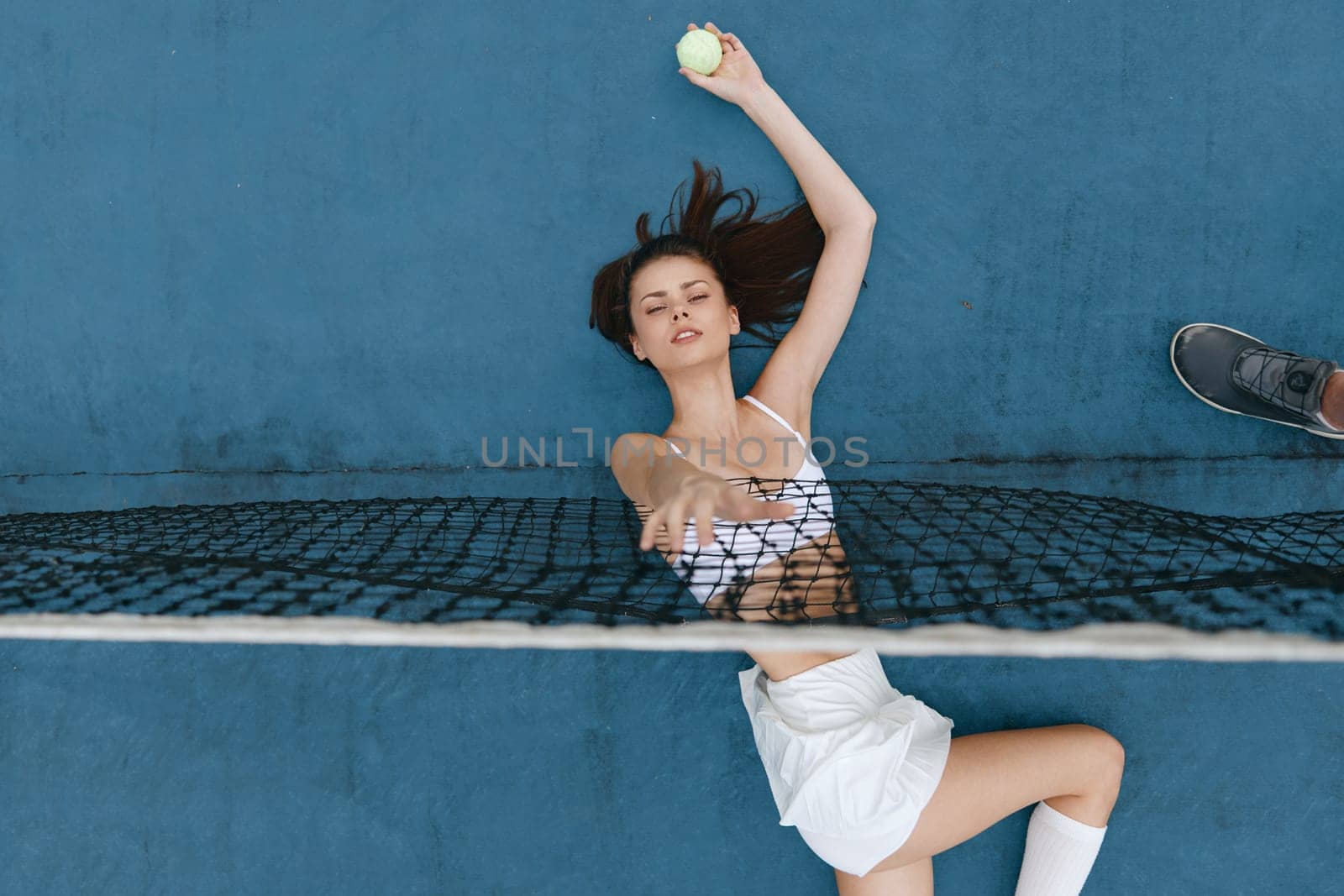A young woman engaging in tennis, radiating confidence and energy while poised to serve The vibrant blue court background highlights her athletic attire and dedication to an active lifestyle