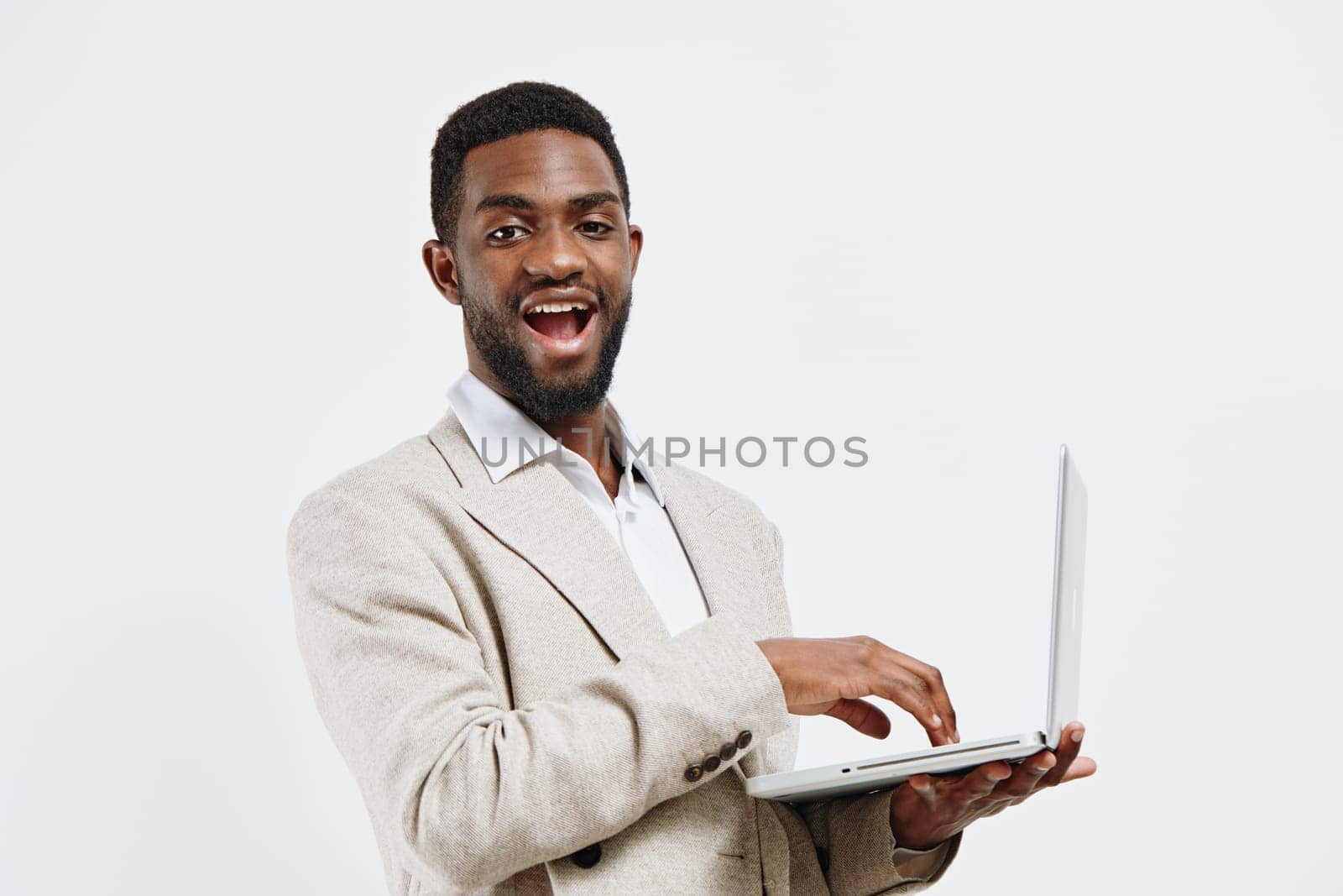 Happy man with a friendly expression holding a laptop computer, dressed in a beige blazer and white shirt, standing against a white studio background, conveying professionalism, confidence, and success, concept: technology and business.