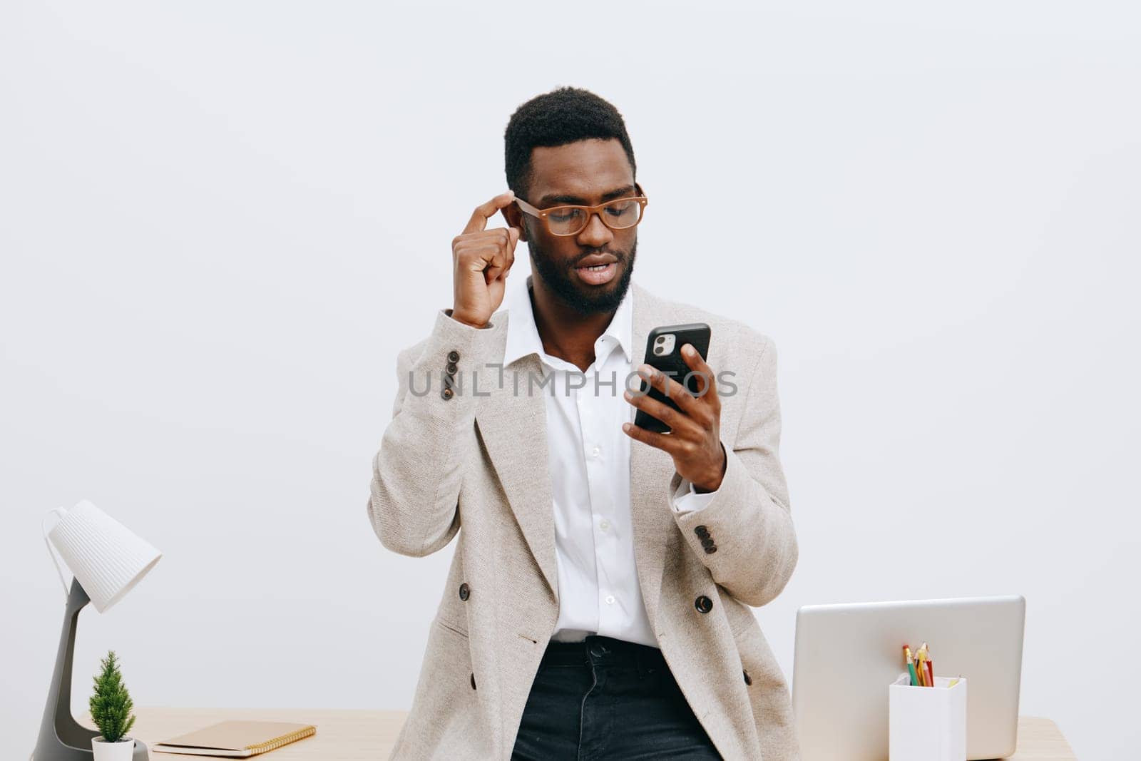 Confident young man wearing glasses and beige blazer using smartphone in minimalist office setting, focused on screen, working remotely or communicating, professional and stylish appearance, clean workspace.