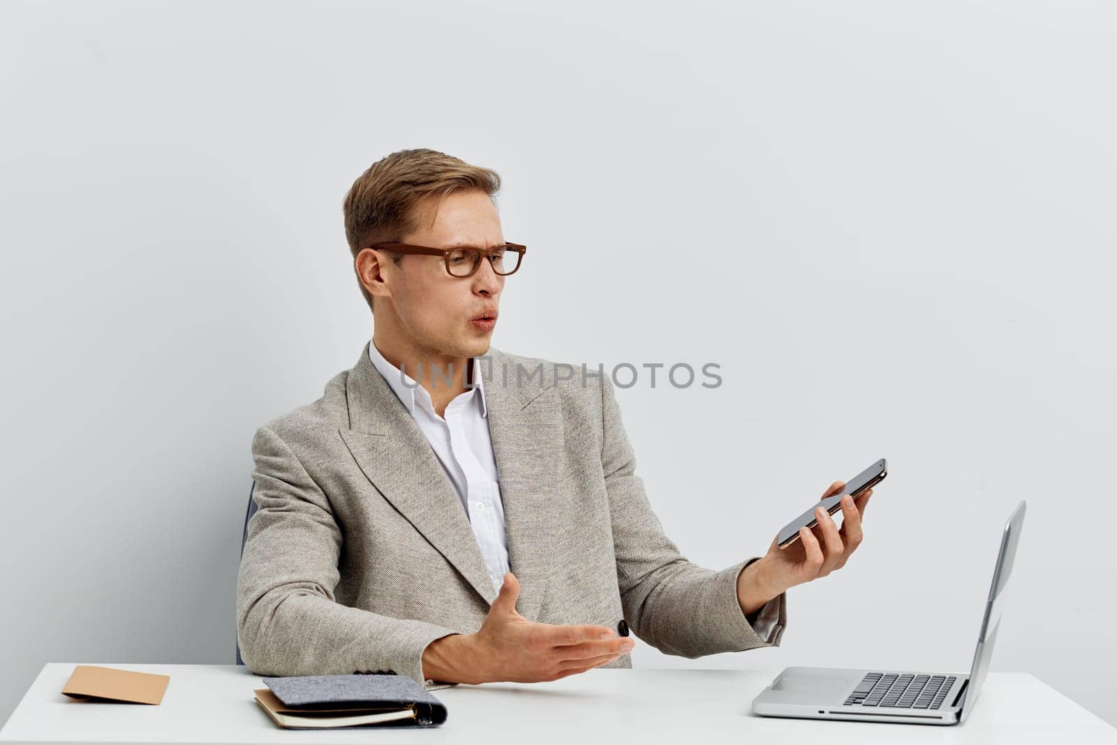 surprised man using smartphone and laptop for digital nomad remote work with glasses and casual jacket on plain white background. He communicates online multitasking with technology gadgets. Concept: digital nomad