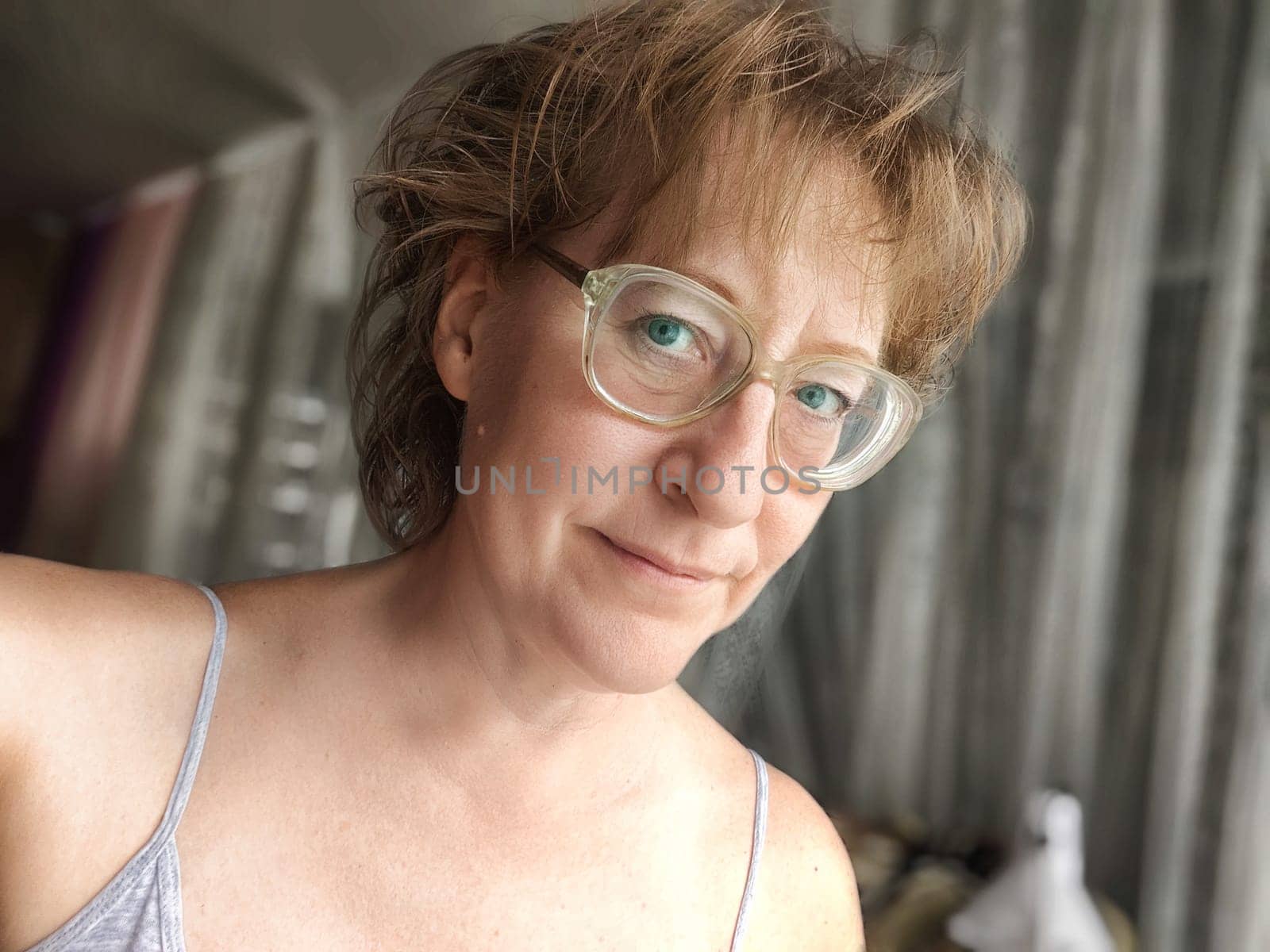 A mature blonde woman smiles in a close-up selfie, capturing her genuine expression and relaxed atmosphere in a cozy indoor setting.