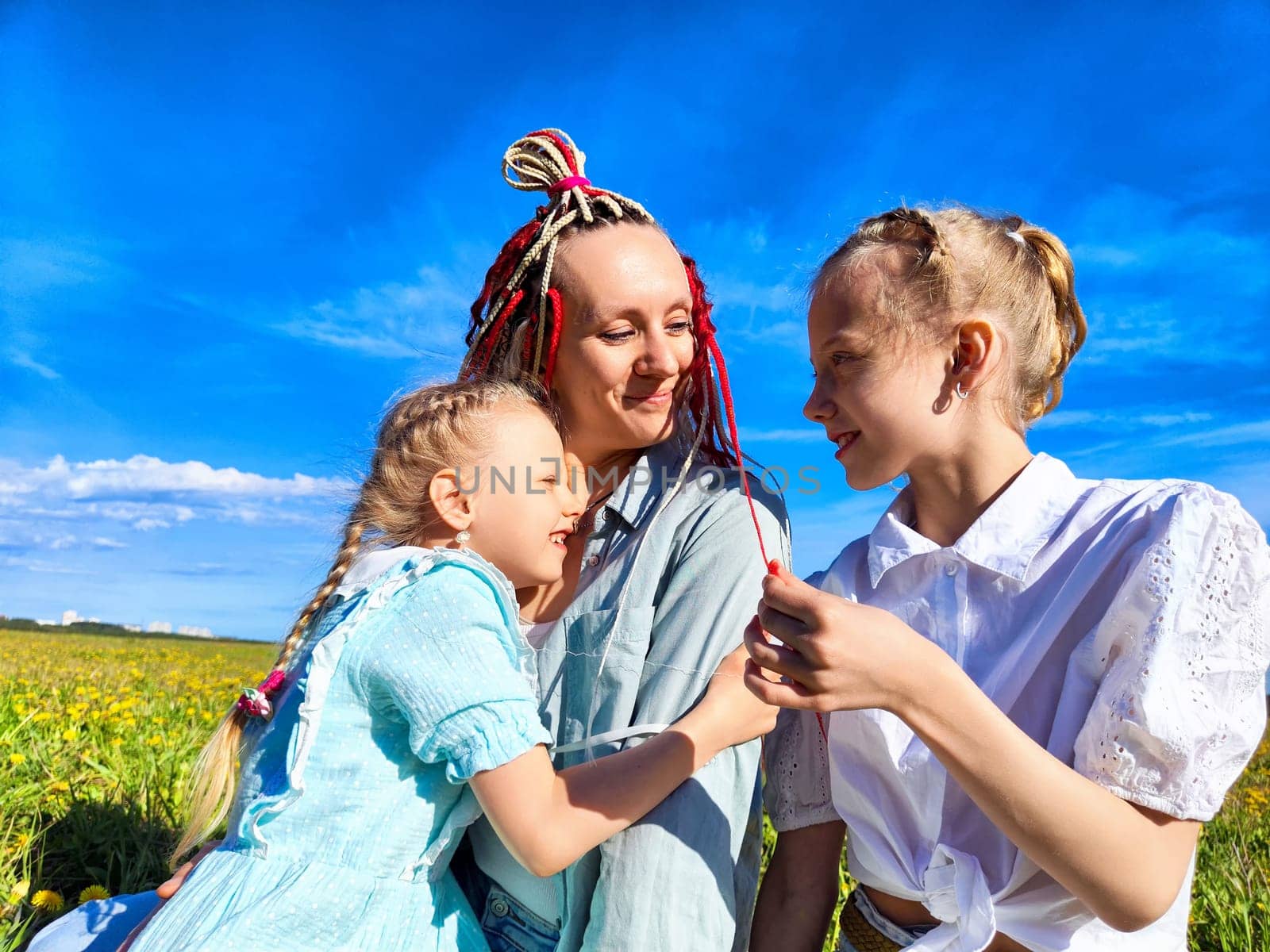 A mother sits with her two daughters in a sunny field, sharing smiles and laughter while surrounded by blooming greenery and bright blue skies.