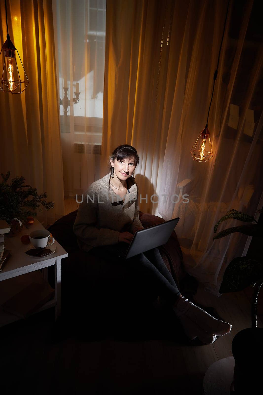 In a warm and inviting apartment, a middle-aged woman relaxes in the evening light, focused on her laptop while sipping a drink.
