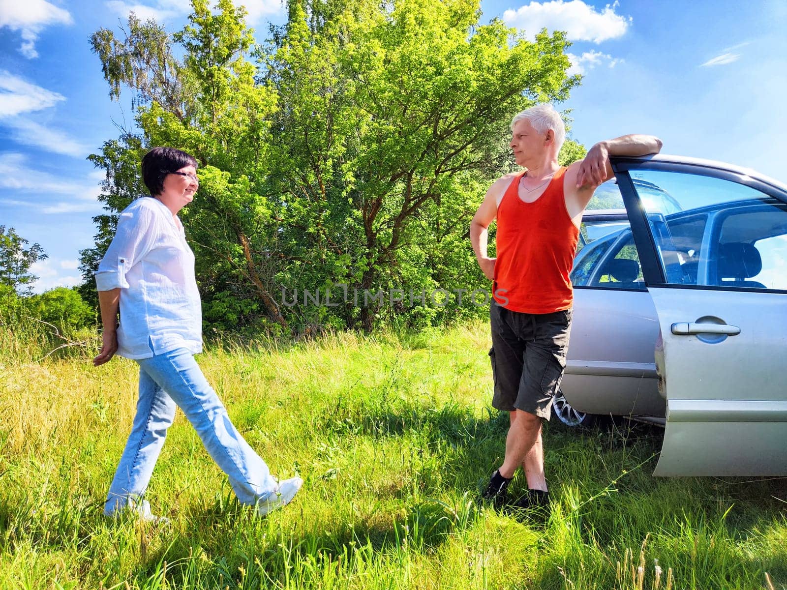 A middle-aged couple stands in a grassy field beside their car, smiling and engaging in conversation. They enjoy a beautiful day outdoors, exploring together and embracing their adventure.