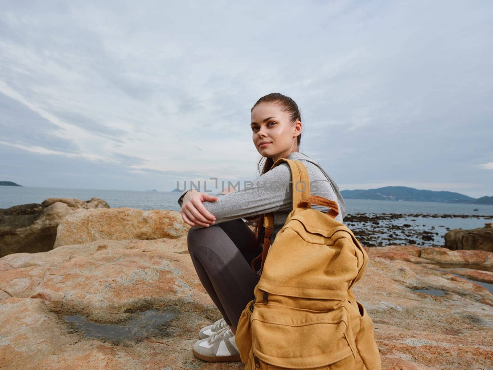 A young woman with a yellow backpack is crouching on rocky terrain by the sea, dressed in casual clothing, showcasing a reflective and thoughtful mood in a serene coastal environment