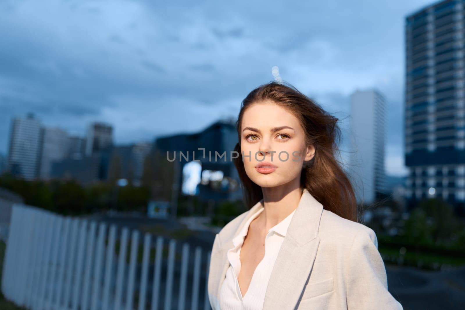 A poised business woman stands confidently in an urban environment, wearing a light-colored suit. The twilight backdrop creates a dynamic atmosphere, emphasizing her professional demeanor.