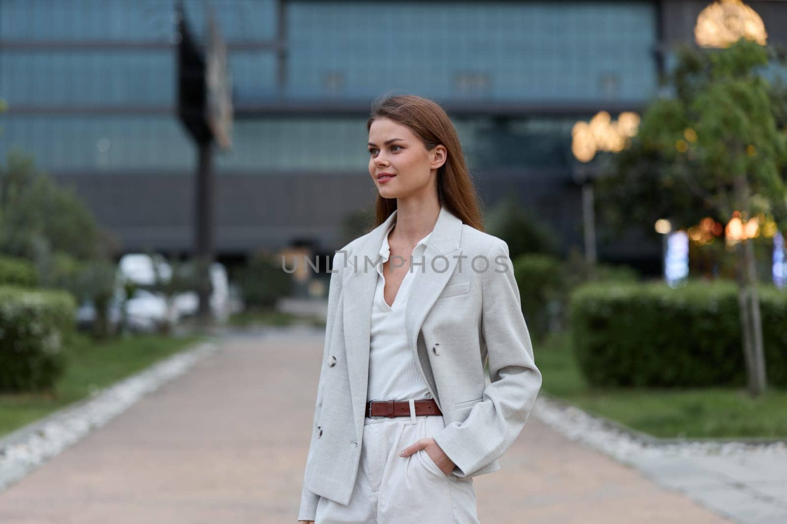 A professional businesswoman in a light-colored blazer and trousers confidently strolls through an urban landscape, exuding poise and sophistication in her demeanor.