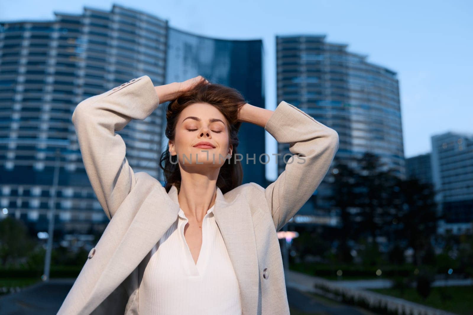 Confident woman in business casual outfit relaxes with eyes closed against urban skyscraper background, expressing peace and tranquility.