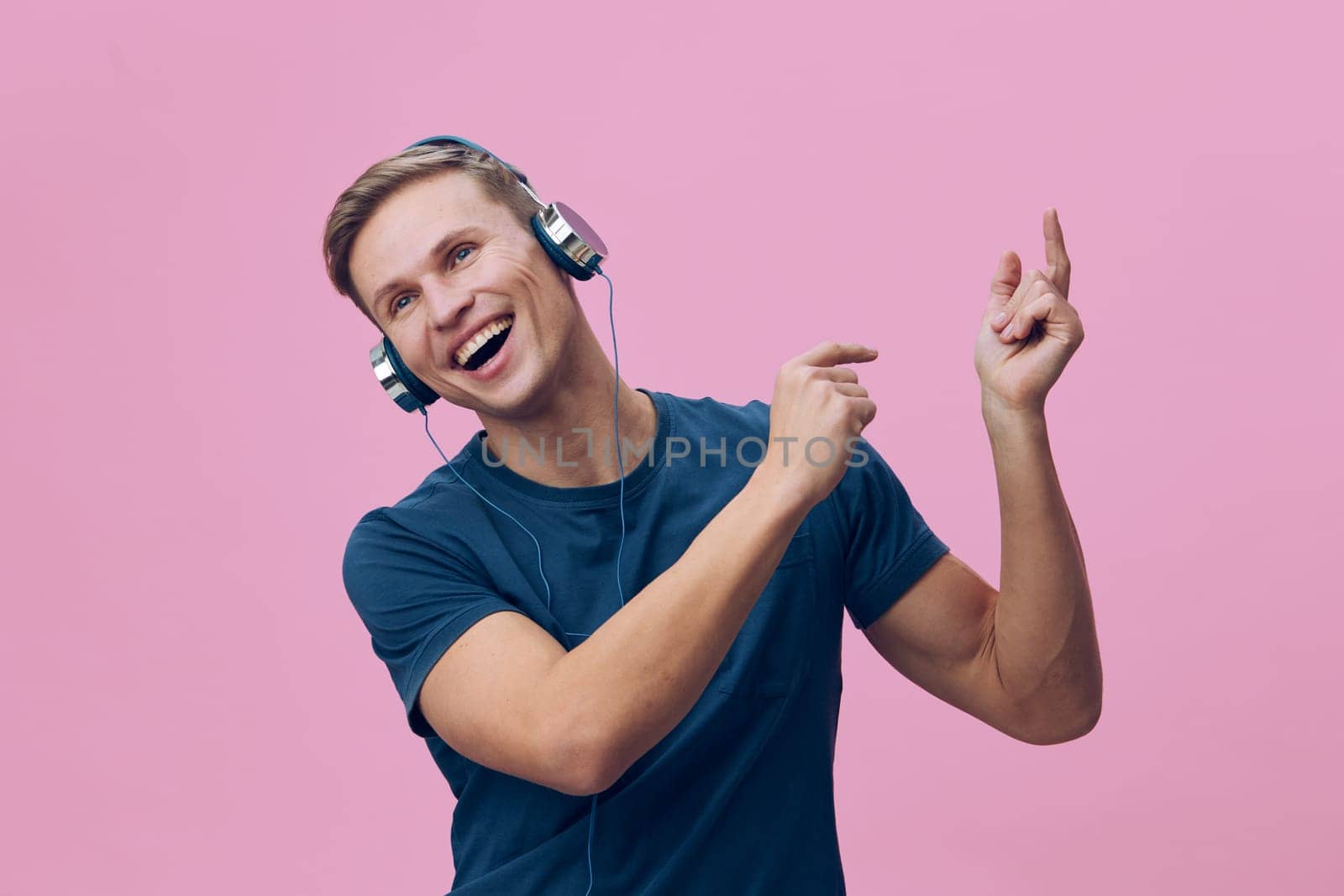 Happy woman wearing headphones expressing joy while dancing, vibrant pink backdrop, casual clothing, youthful energy, music lover, leisure activity, cheerful mood.