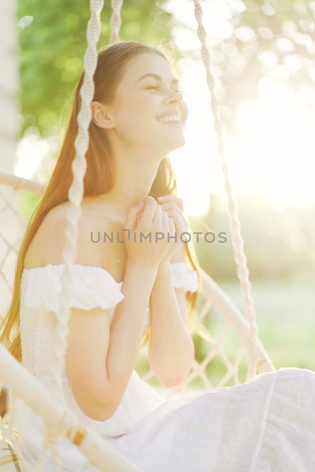 A serene young woman in a white dress smiles while on a swing during a sunny day in a vibrant garden, embodying joy, outdoor happiness, and summer bliss