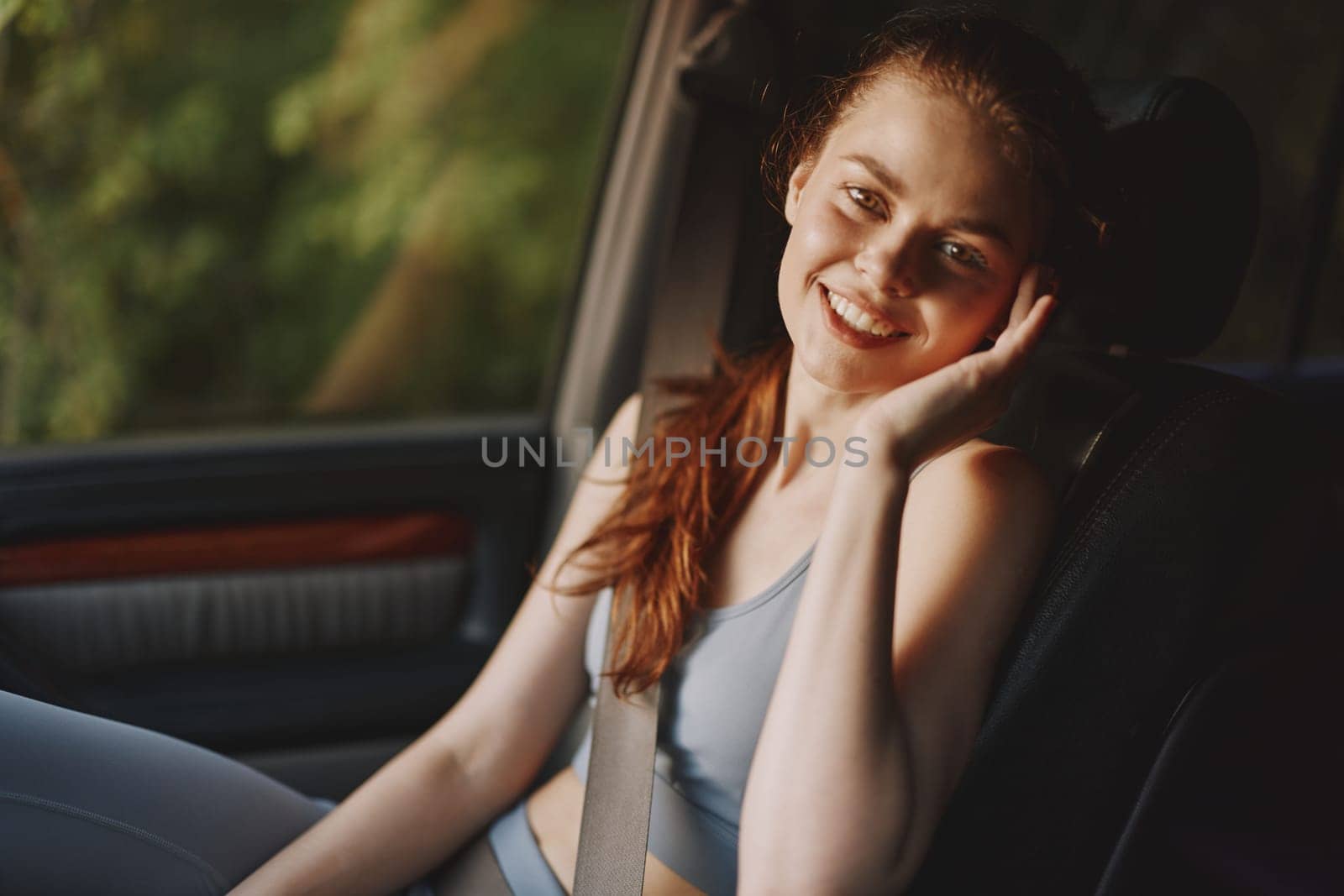 Happy young woman in casual sportswear seated in a car, smiling brightly, emphasizing a joyful and healthy lifestyle reflecting sunny outdoor vibes with natural greenery