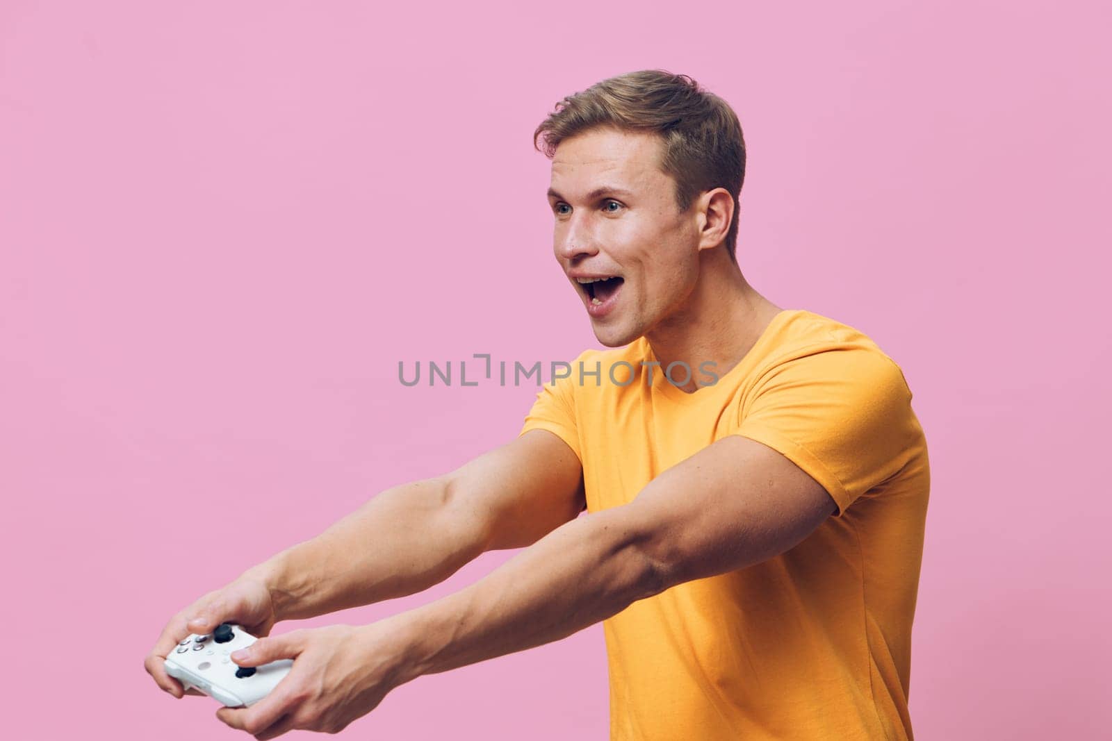 Smiling enthusiastic man having fun gaming, holding game controller with both hands, expressing joy and excitement, dressed in casual orange shirt, pink studio background, lifestyle, leisure, technology concept