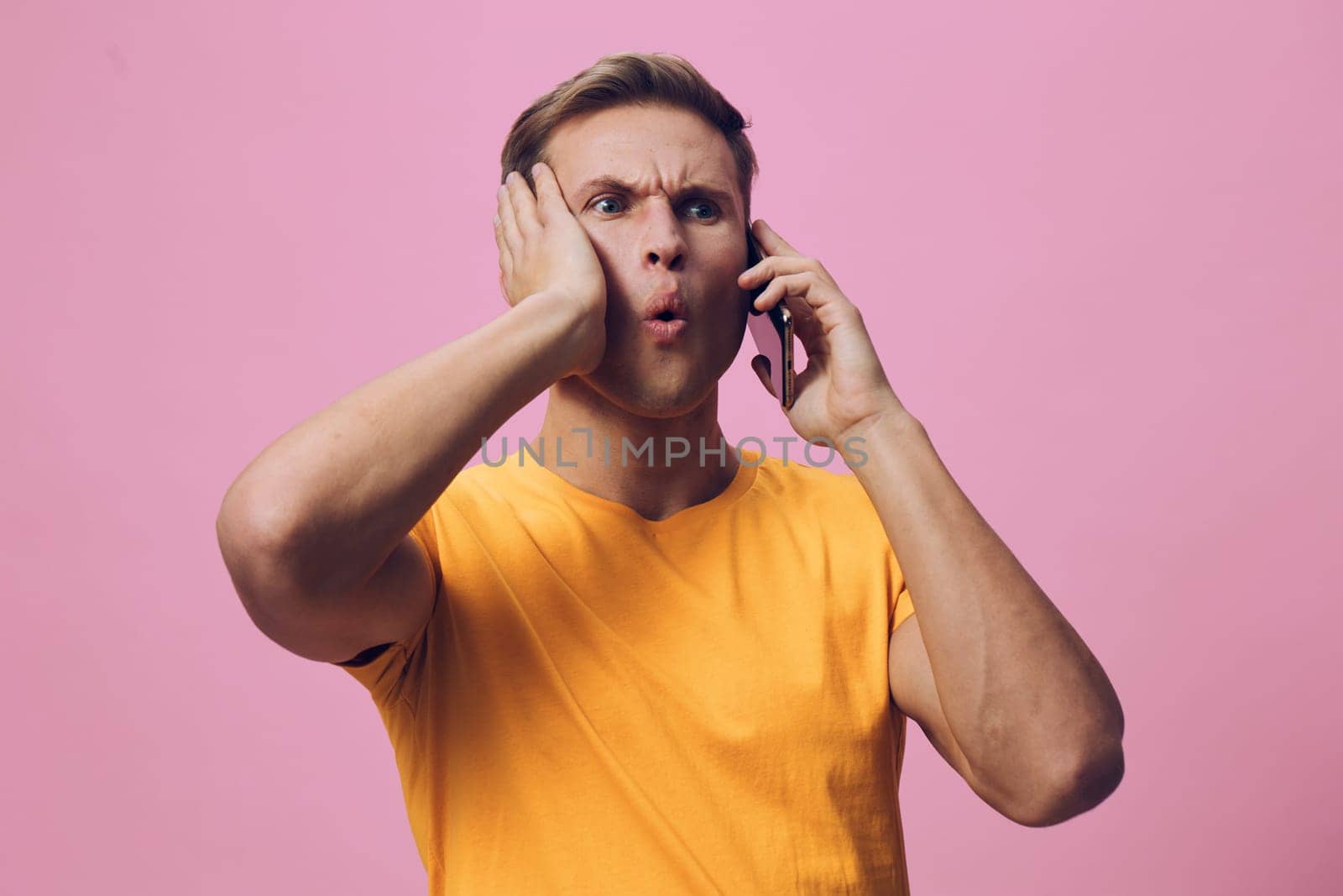 concerned man talking on phone with shocked expression wearing casual orange t-shirt on pink background showing emotions of surprise and confusion digital communication and chat with AI concept