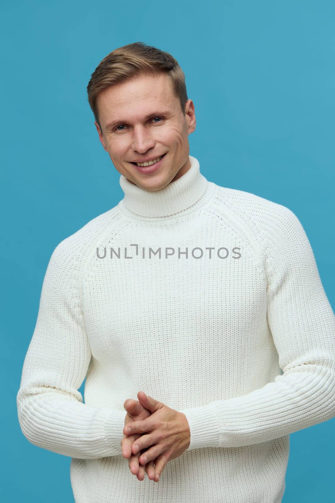 Smiling young caucasian man wearing white knitted turtleneck sweater looks at camera with friendly face while clapping hands on solid blue background studio portrait. Warm casual style and positive emotion concept: mental health and gadgets.