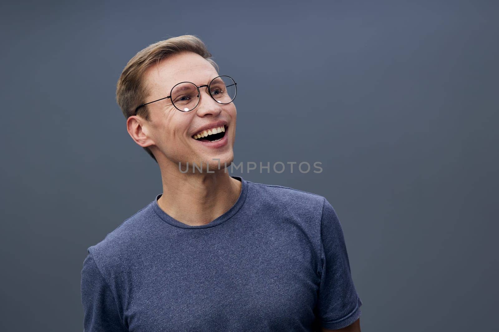 Cheerful young man with glasses and light brown hair wearing casual blue sweater, smiling brightly and looking to the side. Studio shot with plain background, positive vibe, casual style, lifestyle and happiness theme.