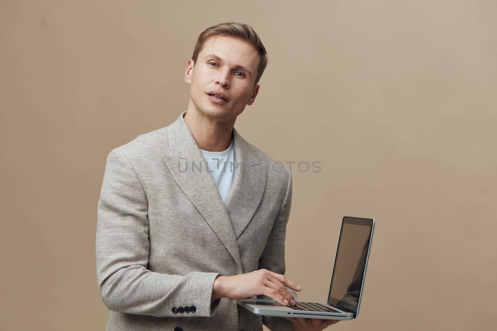 Young handsome caucasian man in smart casual beige jacket hold laptop computer typing browsing internet work remote digital nomad lifestyle on plain background studio shot. Concept: digital nomad work