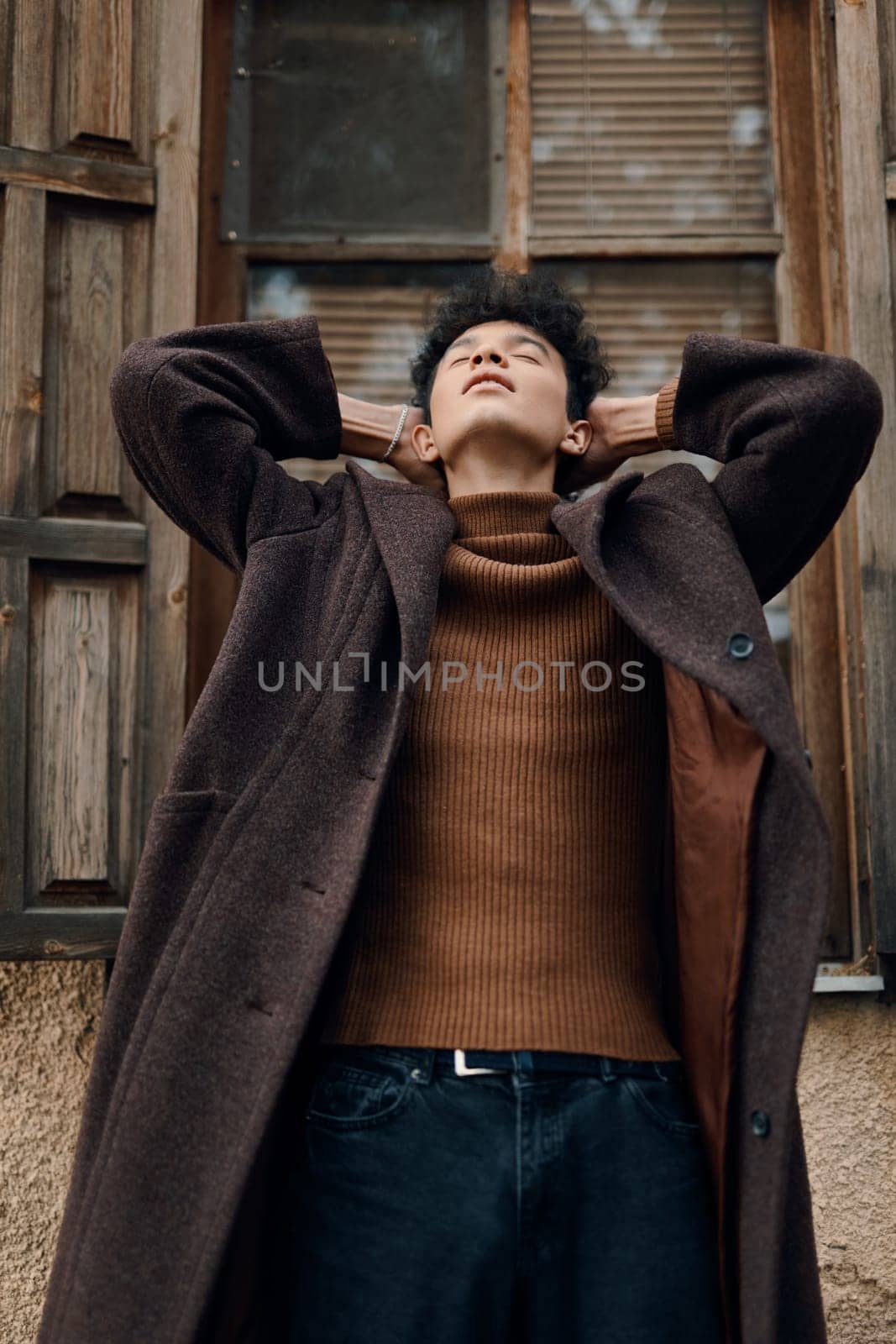 Fashion man in brown coat and turtle neck sweater standing with eyes closed and hands on head in front of window.