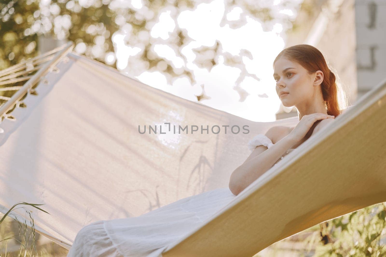 A peaceful young woman in a flowing white dress lounges on a hammock, basking in soft sunlight and surrounded by greenery, evoking a sense of serenity and relaxation